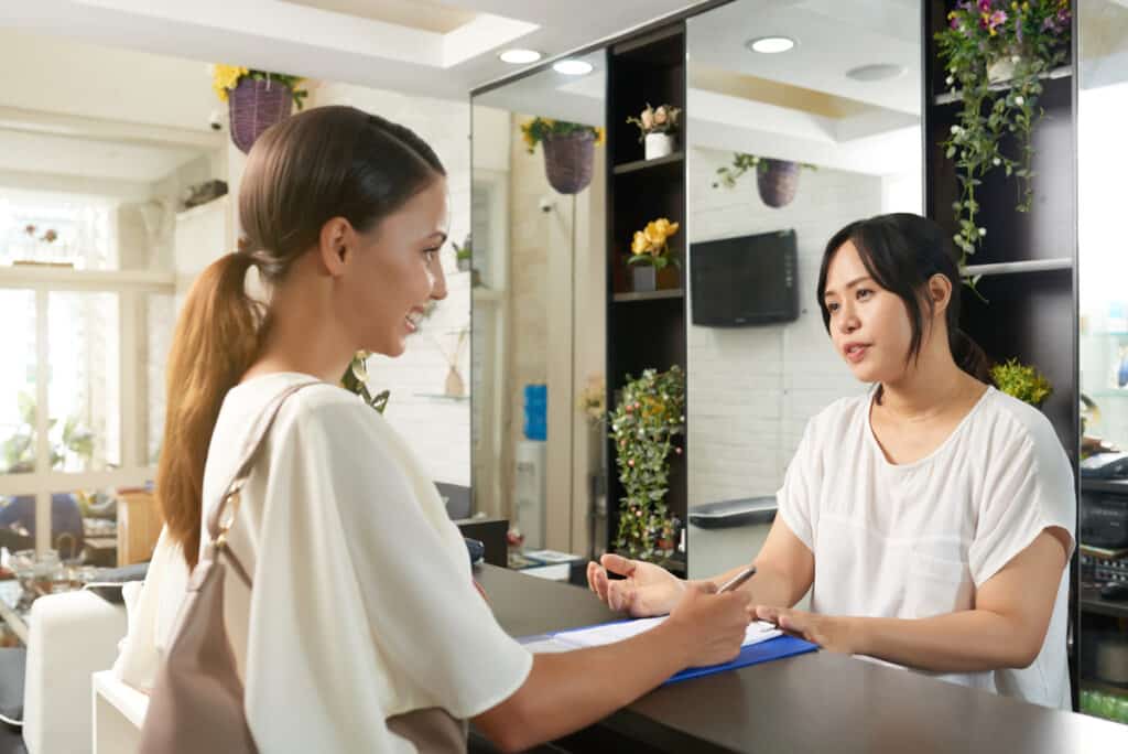 Asian young woman standing near the reception talking to Asian receptionist and filling form before spa procedure in salon