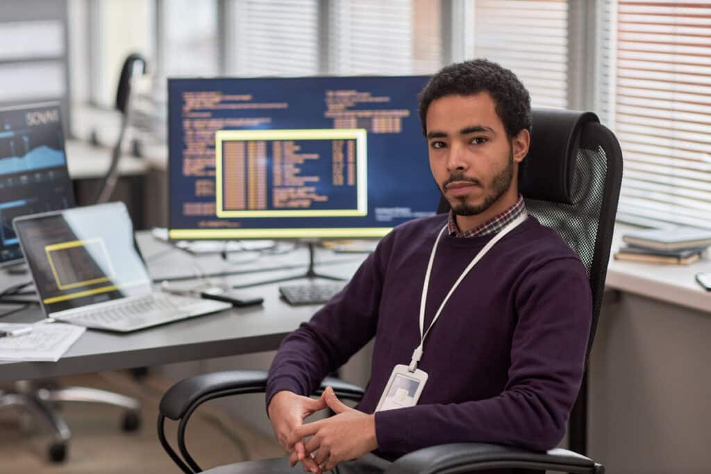 Portrait of male data scientist looking at camera while sitting at desk in IT development office with computers in background